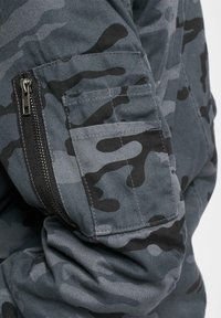 Mørkegrå camouflagejakke med detaljer som en lynlåslomme og tekstureret stof. Designet inkluderer varierende sorte mønstre på overfladen.