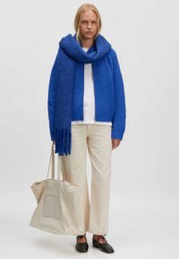 Blauwe gebreide cardigan met bijpassende oversized sjaal, gecombineerd met een witte top, crèmewitte broek en een beige handtas. Zwarte platte schoenen.