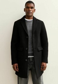 Zwarte wollen blend jas met een gegoten kraag, knoopsluiting en zijzakken, gedragen over een grijze trui en donkere jeans.