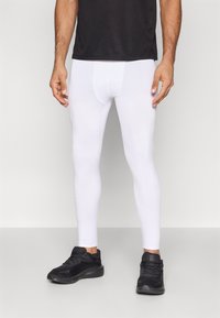 Férfi fehér kompressziós leggingsben és fekete futócipőben, sima háttér előtt állva.