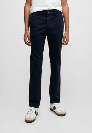 Man draagt marineblauwe rechte broek en witte sneakers met zwarte en beige accenten, staand tegen een effen achtergrond.