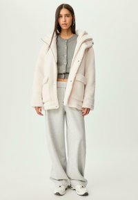 Femme portant une veste blanche en fourrure, un pull gris court avec des boutons, un pantalon de survêtement gris à jambes larges et des baskets blanches, se tenant devant un fond uni.