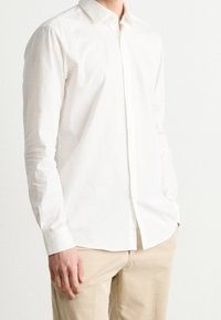 Camicia bianca a maniche lunghe in tessuto leggero con colletto button-down, caratterizzata da un orlo dritto e una texture sottile. Indossata con pantaloni beige.