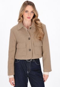 Giacca in lana beige tagliata con due tasche frontali, grandi bottoni e colletto a punta, abbinata a pantaloni in denim scuro.