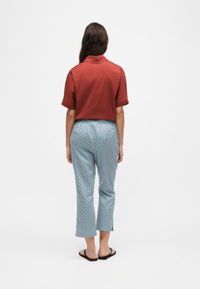 Femme debout de dos, portant une chemise à manches courtes couleur rouille, un pantalon bleu et blanc à motifs géométriques, et des sandales plates noires.