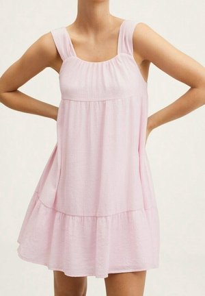 Robe sans manches rose clair en coupe trapèze avec ourlet à volants, encolure carrée et larges bretelles portée par une personne les mains sur les hanches.