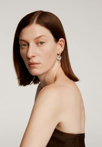 Pendiente de aro dorado con acentos en turquesa; el modelo tiene el cabello lacio y hasta los hombros de color castaño; tono de piel clara; lleva puesto un top marrón sin tirantes.