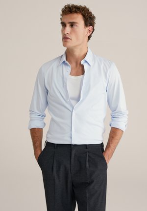 Chemise bleu clair à boutons avec manches retroussées, assortie à un pantalon gris foncé sur mesure. La chemise présente une texture douce et un col classique.