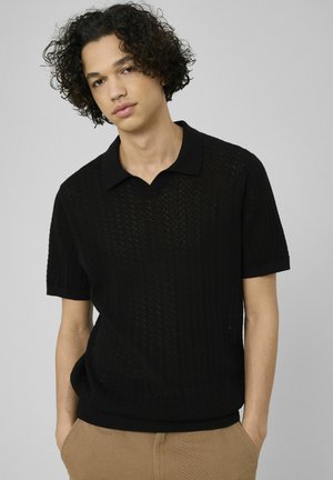 Jeune personne aux cheveux bouclés portant un polo noir tricoté à manches courtes et un pantalon beige, debout devant un fond gris uni.