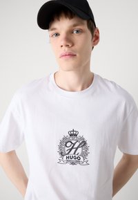 Baltas kokvilnas T-krekls ar melnu grafisko logo, kas ietver sarežģītu dizainu, kroni un tekstu "HUGO SCHOOL OF LIFE" uz krūtīm.