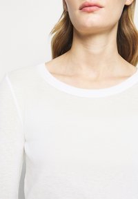 Leichtes, weißes Langarmoberteil mit rundem Halsausschnitt und subtiler Struktur. Verfügt über einen kontrastierenden weißen Saum entlang des Kragens.