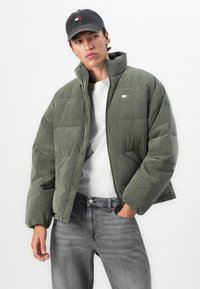 Jeune homme portant une veste en velours côtelé rembourrée verte, une chemise blanche, un jean gris et une casquette de baseball noire, avec une main dans la poche.