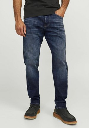 Jeans din denim albastru închis, cu o croială dreaptă, textură ușor decolorată, design cu cinci buzunare, asortați cu pantofi închiși, având o talpă bej deschis.