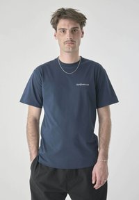 Man with short brown hair and mustache wearing navy blue t-shirt, silver chain necklace, bracelets, and black pants against plain background.