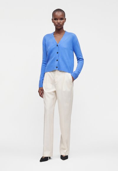 Femme portant un cardigan bleu boutonné, un pantalon crème taille haute et des chaussures noires pointues, debout avec une main dans la poche.