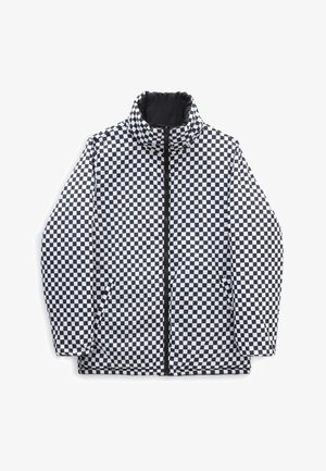 Persona vestida con una chaqueta acolchada negra de Vans con bolsillos laterales y pantalones marrones, de pie contra un fondo blanco.