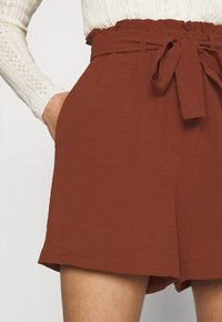 Shorts de couleur rouille fabriqués dans un tissu texturé, avec une large ceinture à nouer et des poches latérales.