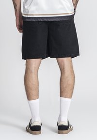 Shorts en denim noir avec une coupe ample, featuring des poches arrière et un ourlet simple. Portés avec des chaussettes blanches et des baskets noir et blanc.
