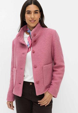 Kvinde iført en lyserød uldjakke med knapper, hvid skjorte, blå og pink halstørklæde og mørke bukser, stående mod en hvid baggrund.