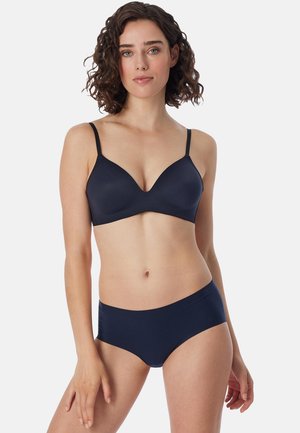 Femme aux cheveux bouclés et foncés, portant un soutien-gorge bleu foncé assorti et un sous-vêtement hipster sans coutures, debout sur un fond clair uni.