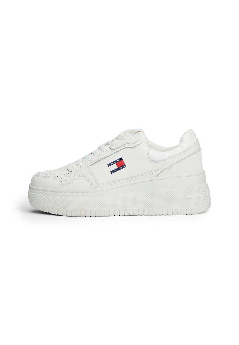 Sneaker en cuir blanc avec une semelle plate, dotée de perforations, d'un logo de marque en rouge et bleu, et d'une semelle en caoutchouc texturée.