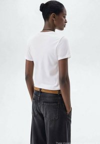 Bílý krátký rukávový crop top v kombinaci s tmavými džíny s vysokým pasem. Mezi pozoruhodné prvky patří hnědý pásek s jemným zdobením.