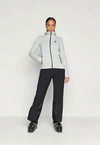 Ανοιχτό γκρι ζακέτα με φερμουάρ και μαύρες λεπτομέρειες και τσέπες, συνδυασμένη με μαύρα παντελόνια και μποτάκια σκι. Λείο υλικό και εφαρμοστή σχεδίαση.