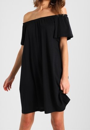 Femme portant une robe noire ample, épaules dénudées, tenant l'ourlet d'une main devant un fond clair uni.