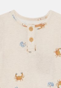 Beige bomullsskjorte med rund hals, med små krabbe- og blå blomstertrykk, og to knappelukkinger i tre ved halsen.