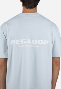 Svetlo modra bombažna majica s kratkimi rokavi, na hrbtni strani je bel tiskani grafični motiv z napisom "PEGADOR" in pod njim "CERTIFIED AUTHENTIC STREETWEAR".