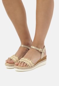 Sandalias de cuña con parte superior de paja trenzada, correa para el tobillo de cuero beige y plataforma de corcho texturizado. Suela de goma blanca con detalles de trenzado.