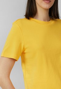 T-shirt en coton jaune à manches courtes, col rond et texture lisse. Un léger détail aux épaules améliore le design.