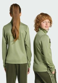 Dvaja jednotlivci oblečený v zladených olivovozelených športových mikinách na zips a nohaviciach. Tkanina vyzerá hladko s jemným leskom.