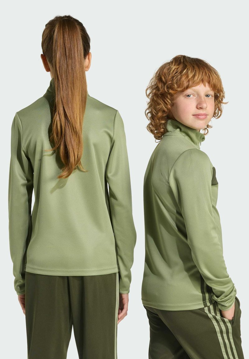 Dvaja jednotlivci oblečený v zladených olivovozelených športových mikinách na zips a nohaviciach. Tkanina vyzerá hladko s jemným leskom.