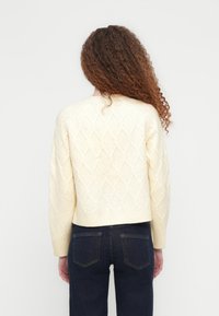 Γυναίκα με μακριά σγουρά μαλλιά, φορά ει μάλλινο πουλόβερ σε εκρού χρώμα με διαμάντι μοτίβο και σκούρο μπλε τζιν, στέκεται μπροστά σε απλό φωτεινό φόντο.