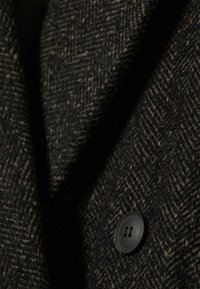 Houndstooth-patterned wool fabric in black and brown tones, featuring a large black button, detailed texture visible in close-up.