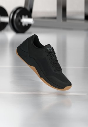 Chaussure de sport noire avec semelle en gomme flottant au-dessus du sol de la salle de sport, barre d’haltérophilie floue en arrière-plan.