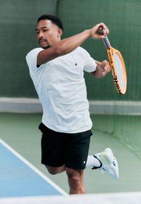 Tennisspieler in einem kurzärmeligen weißen Shirt und schwarzen Shorts, der mit einem orangefarbenen Tennisschläger auf einem grünen Platz mit glatter Oberfläche schwingt.