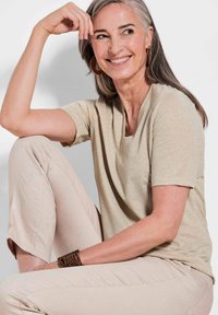 Beige T-shirt met korte mouwen en een ronde hals, gecombineerd met lichtbeige gebloemde broek. De outfit heeft een subtiele glans en een gestructureerde armband.