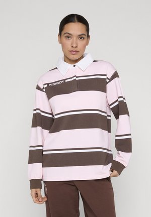 Kvinde iført en langærmet rugbytrøje med pink og brune striber og hvid krave, stående mod en ensartet lys baggrund.