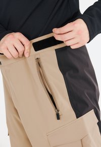 Beige och svarta cargo-byxor med en ficka med dragkedja, justerbar velcro midja och skräddarsydd passform. Mjuk, flexibel tygstruktur.