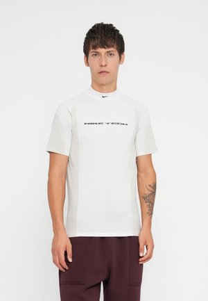 Hvid kortærmet skjorte lavet af letvægtsstof, med høj krave og 'NIKE TECH' logo i mørkegrå over brystet.