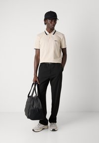 Beżowa koszulka polo z czarnym kołnierzykiem, czarne spodnie, szare sneakersy i czarna torba na ramię, wyeksponowane na jednolitym szarym tle.
