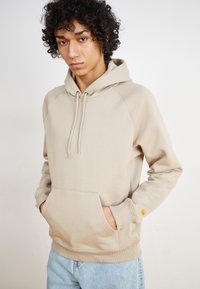Beige hooded sweatshirt with ribbed cuffs, drawstring closure, and front pocket. Small logo detail on sleeve. Soft, textured fabric.