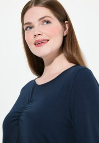 Top blu navy realizzato in morbido tessuto, con scollatura rotonda e dettagli arricciati al centro davanti. Tessuto liscio e vestibilità rilassata.