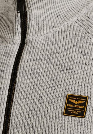 Pull en tricot gris et blanc avec un col montant, fermeture éclair intégrale, motif texturé et un patch logo noir et doré sur le côté gauche.