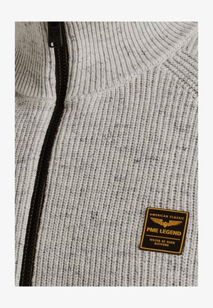 Grauer und weißer Strickpullover mit hohem Kragen, vollem Reißverschluss, strukturiertem Muster und einem schwarz-goldenen Logo-Patch auf der linken Seite.