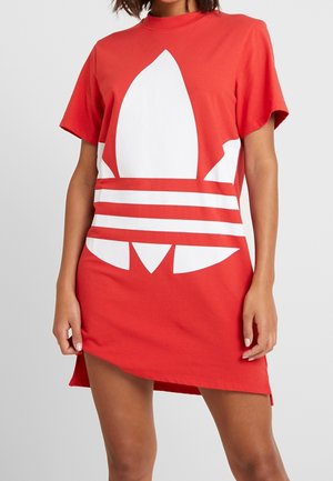 Personne portant une robe t-shirt rouge ample avec un grand logo Adidas trefoil blanc sur le devant, debout devant un fond uni.