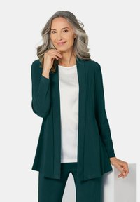 Een lichtgewicht teal cardigan over een witte top met subtiele versieringen, gecombineerd met bijpassende onderstukken, voorzien van lange mouwen en een open voorkant.