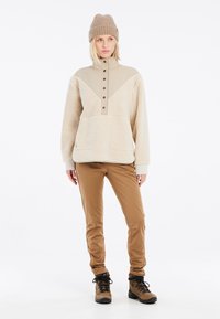 Beige fleece trui met hoge kraag, knoopsluiting en borstzak; gecombineerd met tan slim-fit broeken en bruine wandelschoenen.